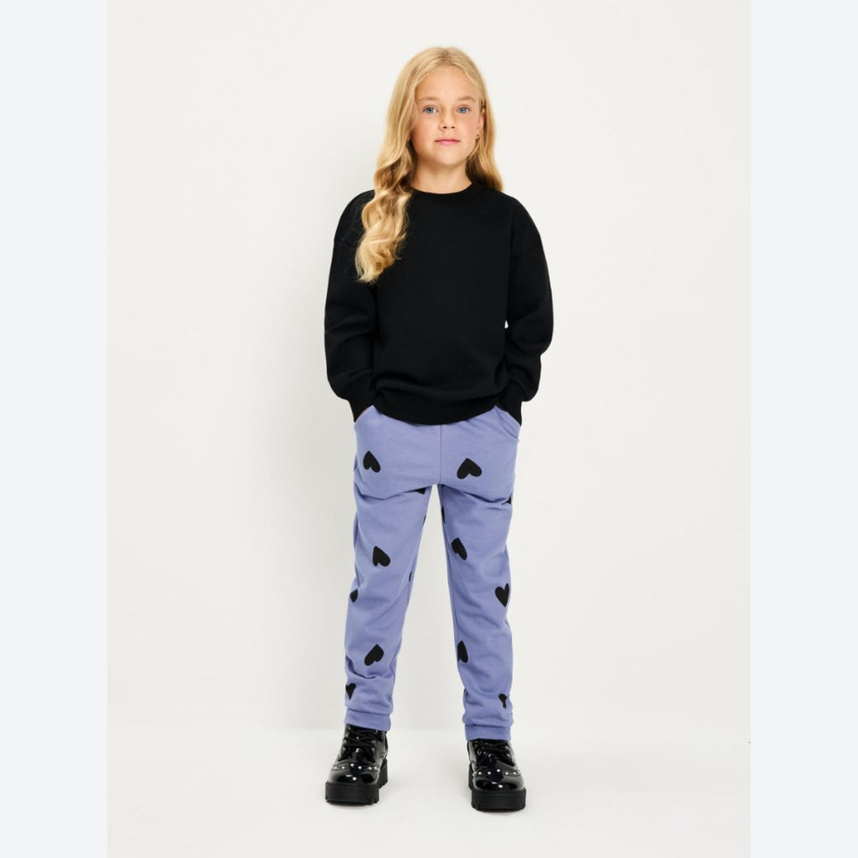 A young girl stands against a plain light background wearing a black crewneck sweatshirt, lavender pants with black heart patterns, and black shoes.