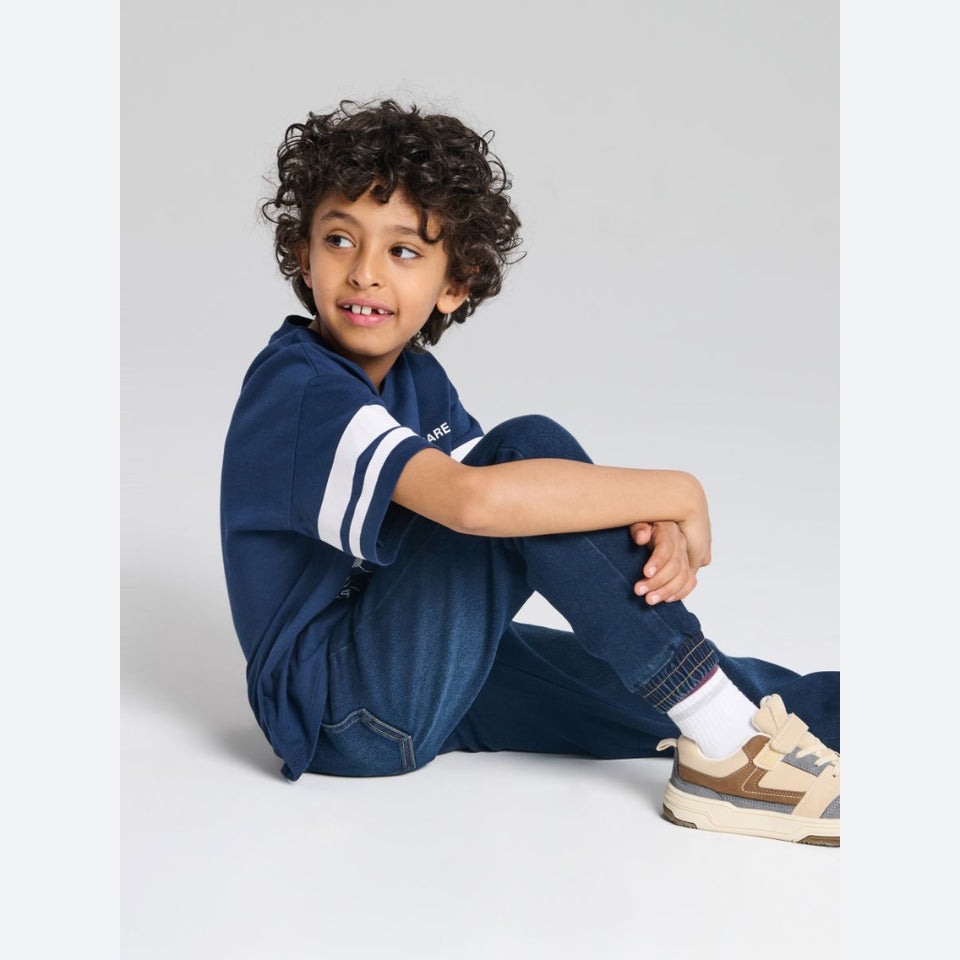 A young boy with curly dark hair sits on a light gray studio floor wearing a navy blue short-sleeve athletic shirt with white stripes on the sleeves, dark blue jeans, white socks, and beige and navy sneakers.