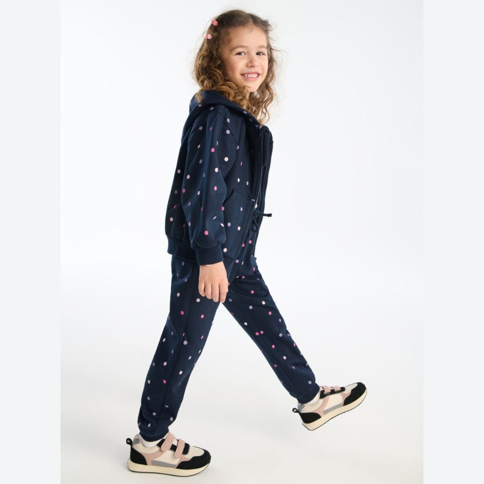 A young girl wearing a navy blue hooded sweatshirt and matching jogger pants, both decorated with a small multicolor polka dot pattern, walking against a plain white studio background.