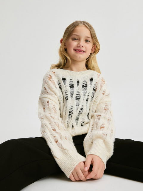 Young girl wearing a white knit sweater sitting on a white surface with a plain background