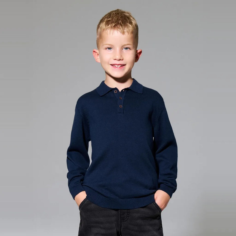 A young boy with blonde hair smiling, wearing a dark navy blue long-sleeve polo sweater and black pants, standing against a solid gray studio background.