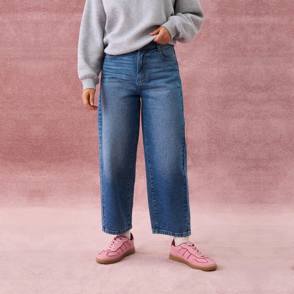 A person wearing blue wide-leg cropped jeans, a light gray sweatshirt, and pink sneakers, standing against a textured pink backdrop.