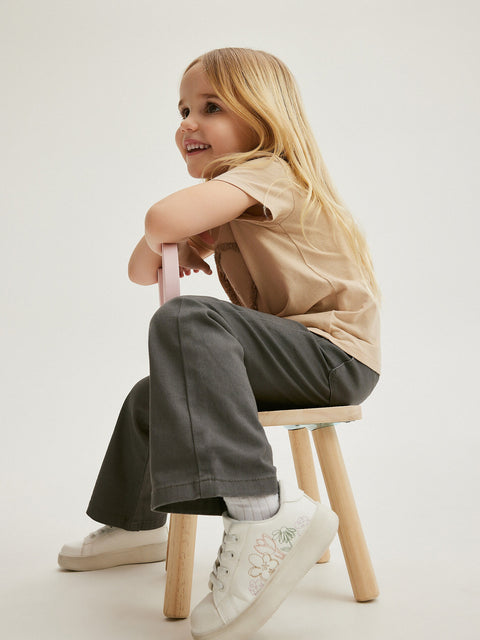 Child sitting on a stool wearing a beige shirt, gray pants, and white sneakers on a plain background