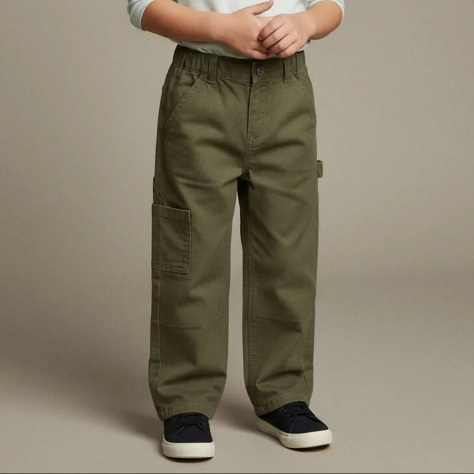 A child wearing olive green cargo pants with side flap pockets, paired with a white shirt and navy blue sneakers, standing against a neutral beige studio background.