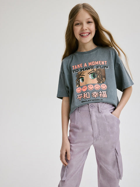 Young girl wearing a gray t-shirt with a graphic design and text, standing against a plain background.