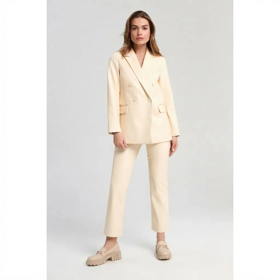 A woman modeling a cream-colored double-breasted blazer and matching cropped trousers with beige loafers, set against a plain light gray studio background.