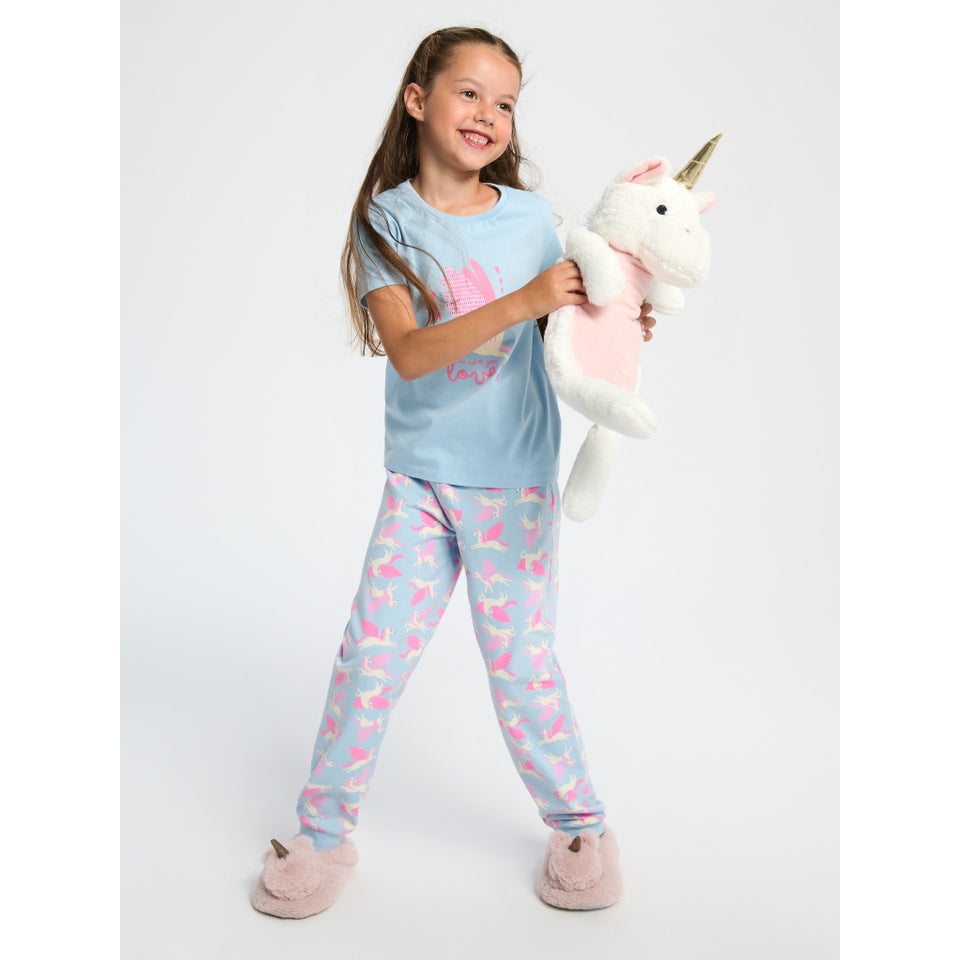A young girl stands against a light gray studio background wearing a light blue pajama set with pink mermaid prints and holding a plush unicorn toy.