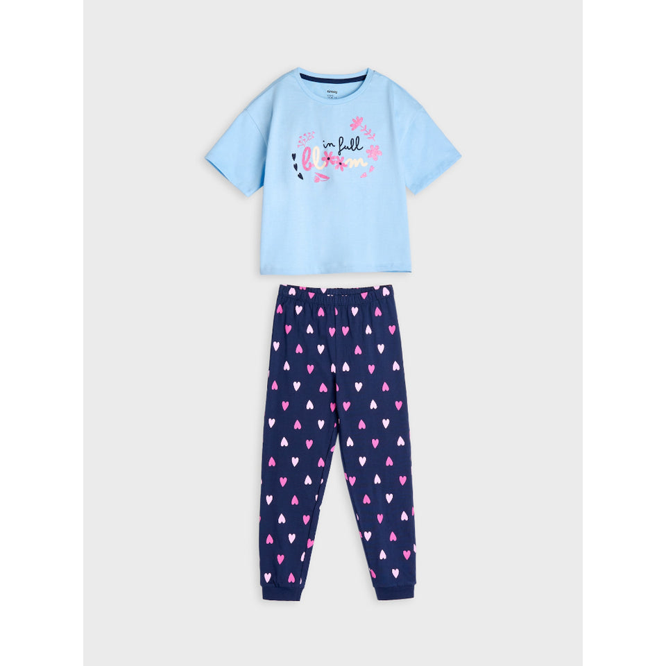 A two-piece children's pajama set laid flat on a light gray background. The set includes a light blue short-sleeve top with a pink and black graphic print on the front, and navy blue jogger pants covered in a pattern of small pink, white, and dark blue hearts.