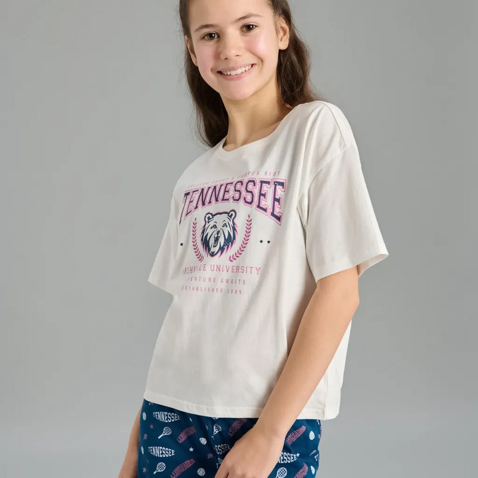 A young girl smiling while wearing a white short-sleeve graphic t-shirt with a purple and blue Tennessee Volunteers logo, paired with blue patterned shorts, against a solid gray studio background.