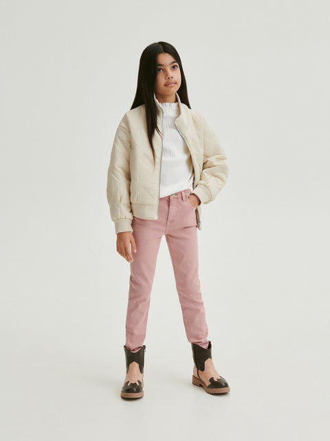 Young girl wearing a cream jacket, white shirt, pink pants, and black shoes on a white background