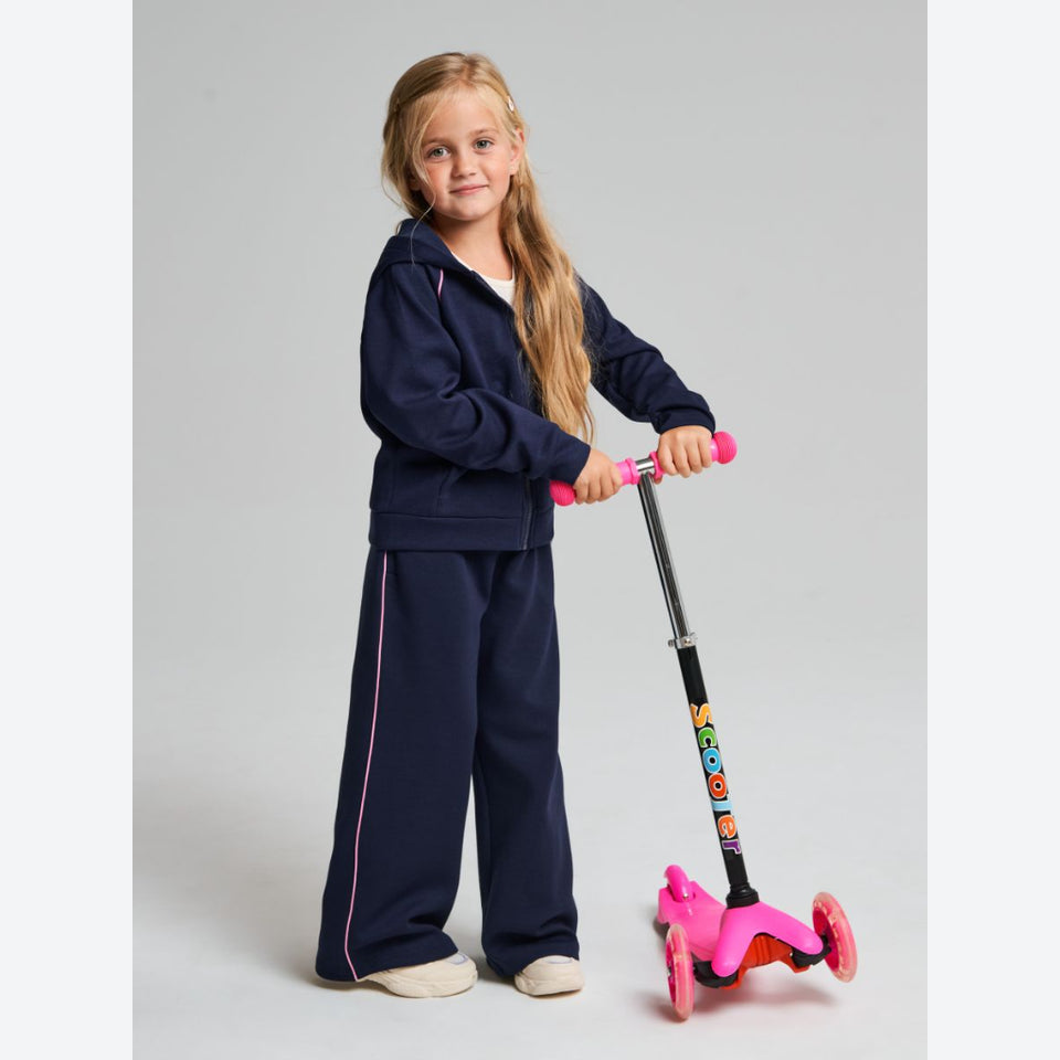 A young girl with long blonde hair stands on a gray studio background, wearing a navy blue tracksuit and holding a pink and black scooter.