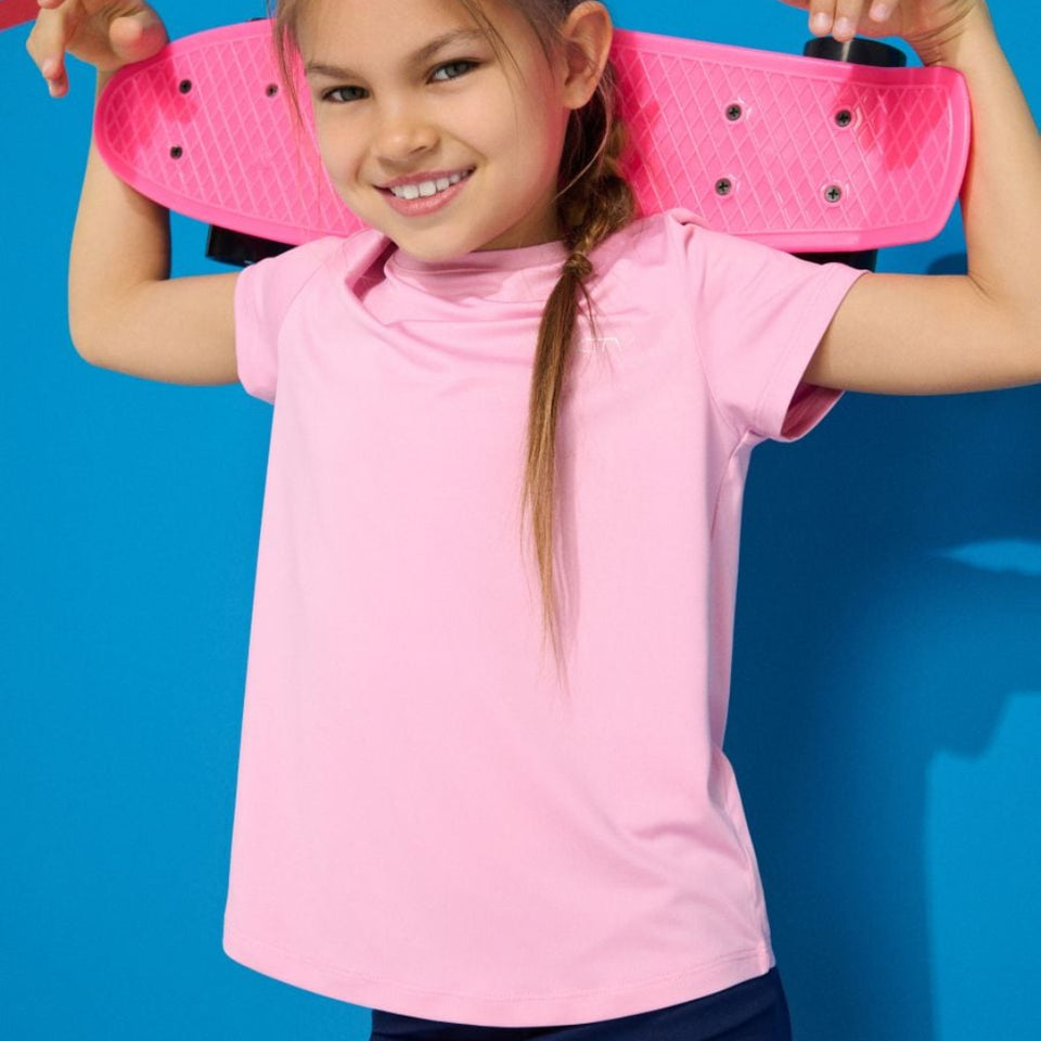 A young girl with a side braid smiles while holding a pink skateboard behind her shoulders, wearing a light pink short-sleeve t-shirt and navy pants, set against a bright blue background.