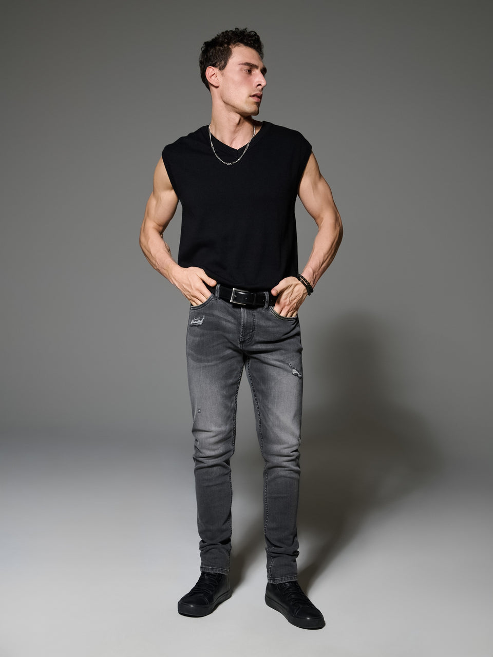 Man wearing a black sleeveless shirt and gray jeans against a gray background