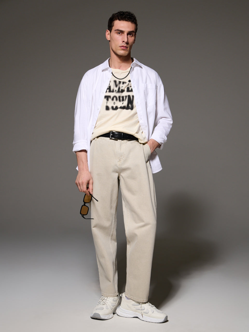 Man wearing a white shirt with text, beige vest, and pants on a gray background