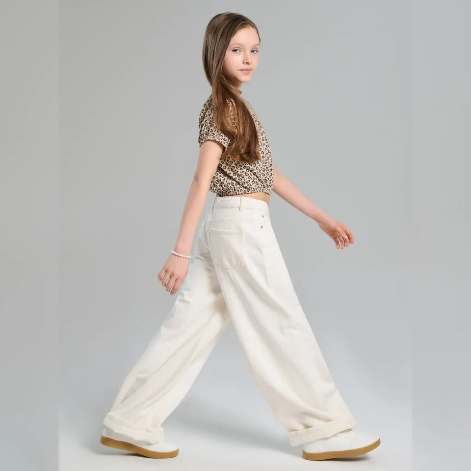 A young girl with long brown hair is shown from a side-back angle, wearing a leopard-print short-sleeve crop top and high-waisted, wide-leg off-white trousers. She is walking against a solid light gray studio background.