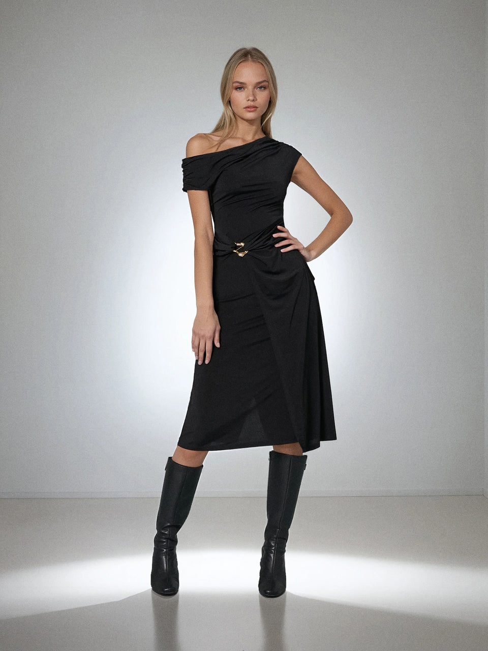 A model wearing a black off-the-shoulder midi dress with a draped waist and a gold-tone belt, paired with black knee-high boots, against a studio backdrop.