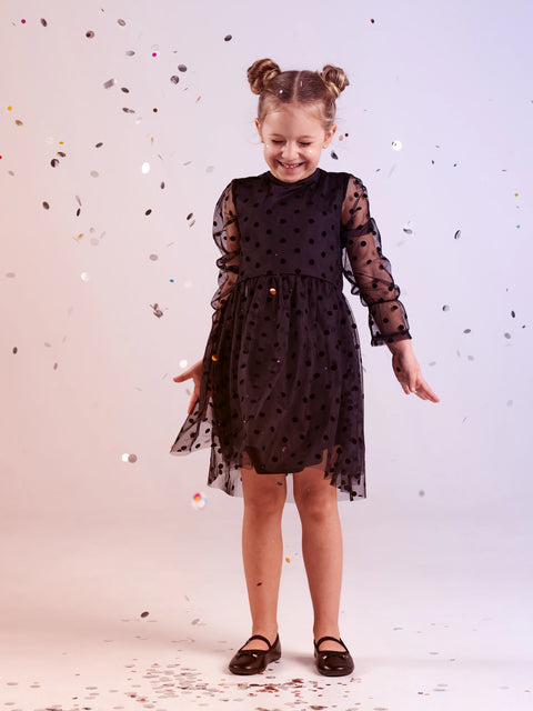 Young girl wearing a black dress with sheer sleeves and polka dot pattern, standing against a light background with confetti.