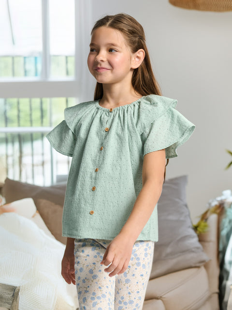 Young girl wearing a light green blouse with ruffled sleeves in a living room setting.