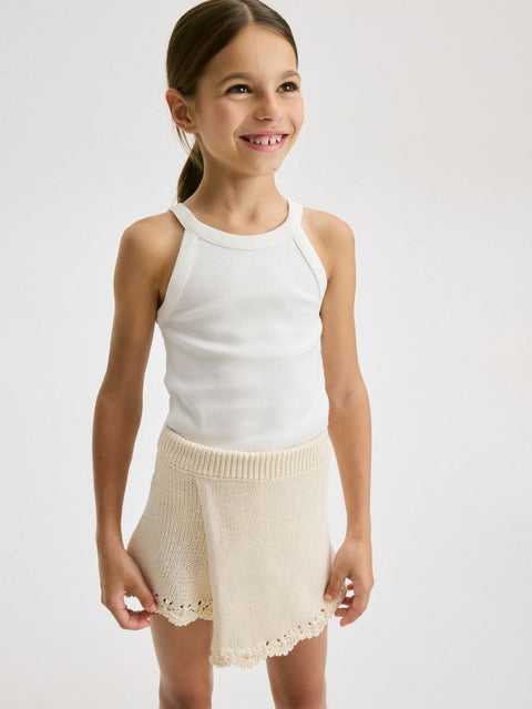 Young girl wearing a white sleeveless top and cream shorts on a light gray background