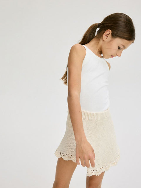 Young girl wearing a white sleeveless top and cream skirt on a plain background
