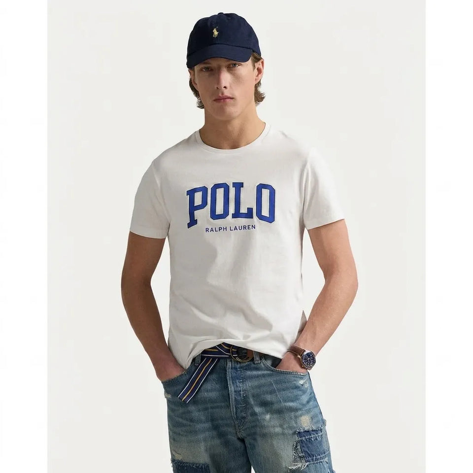 A man wearing a white short-sleeve Polo Ralph Lauren t-shirt with a blue collegiate-style 'POLO' graphic on the chest, paired with distressed blue jeans and a navy blue baseball cap, set against a plain light background.
