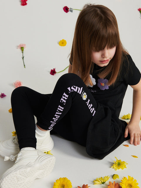 Child sitting on a light-colored floor with flowers around, wearing a black outfit with text and graphics.