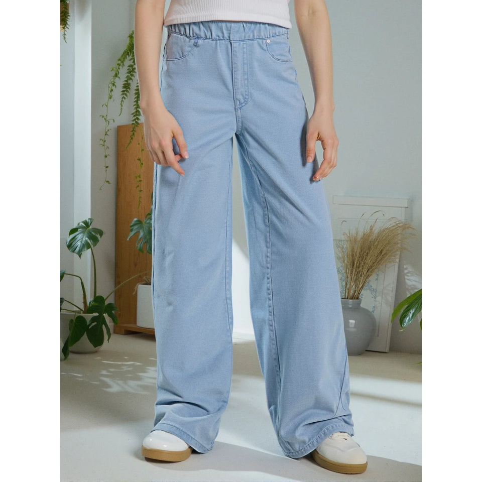 A person wearing light blue wide-leg denim jeans with an elastic waistband, paired with a white top and white sneakers, standing in a bright indoor room with plants and decor.