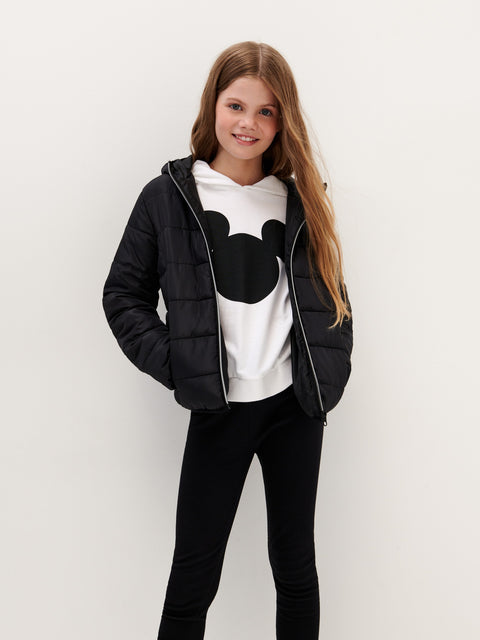 Young girl wearing a black puffer jacket over a white shirt with a black design, standing against a plain background.