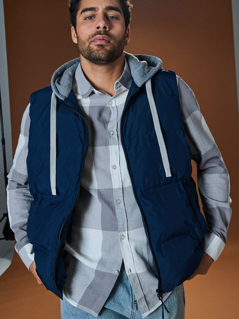 Man wearing a navy blue vest over a plaid shirt with a neutral background