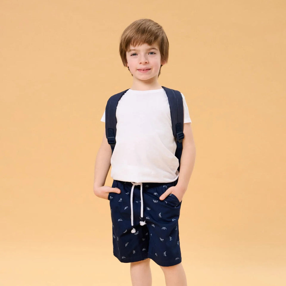 A young boy stands against a solid beige studio background wearing a white raglan t-shirt, navy blue shorts with a small white pattern, a matching navy backpack, and navy sneakers.
