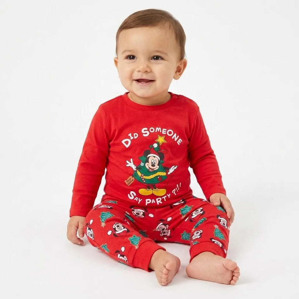 A baby sitting on a white studio background wearing a red long-sleeve pajama top and matching red pajama pants with holiday graphics and text.