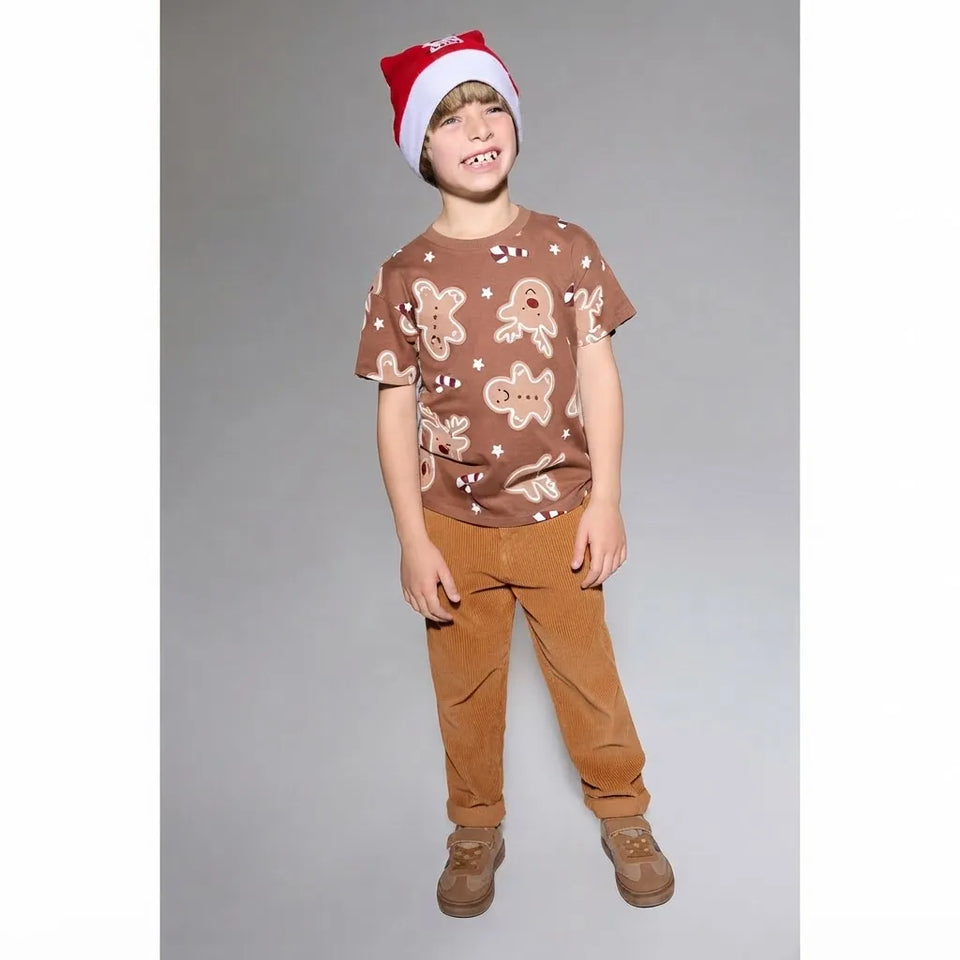 A young boy wearing a brown short-sleeve t-shirt with a gingerbread man pattern, brown pants, and a red and white Santa hat, standing against a solid gray studio background.
