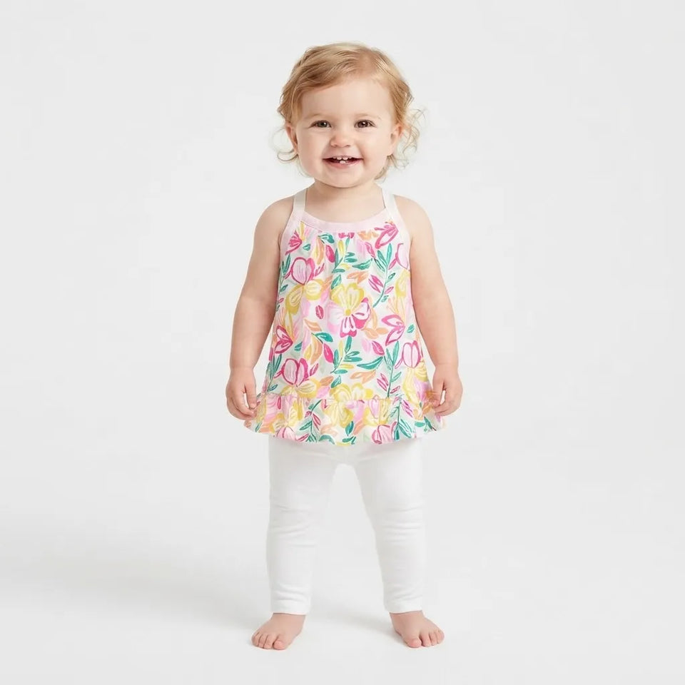 A toddler girl standing barefoot against a plain white studio background, wearing a sleeveless white top with a colorful floral print in pink, yellow, and green, paired with white leggings.
