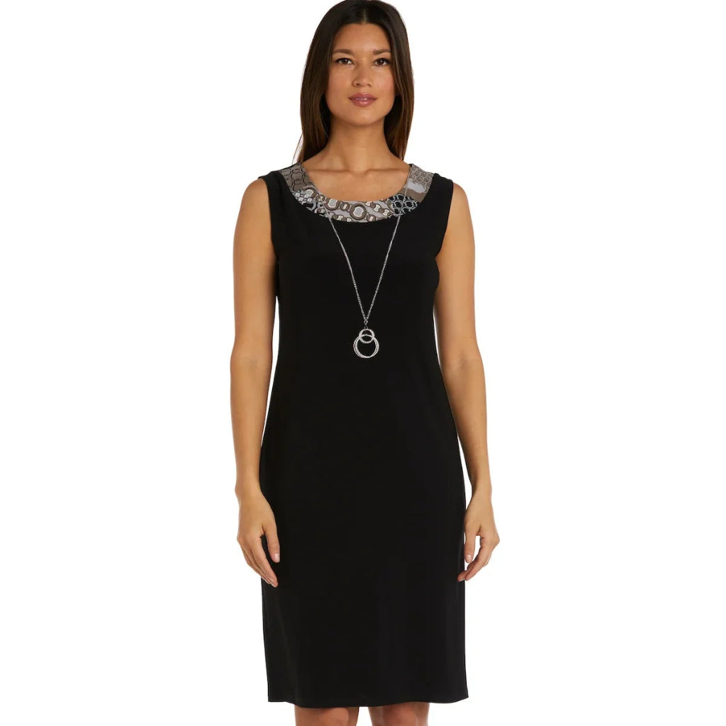 A woman modeling a sleeveless black knee-length dress with a decorative silver and black patterned yoke at the neckline, paired with a long silver pendant necklace and beige heeled sandals, against a plain white background.