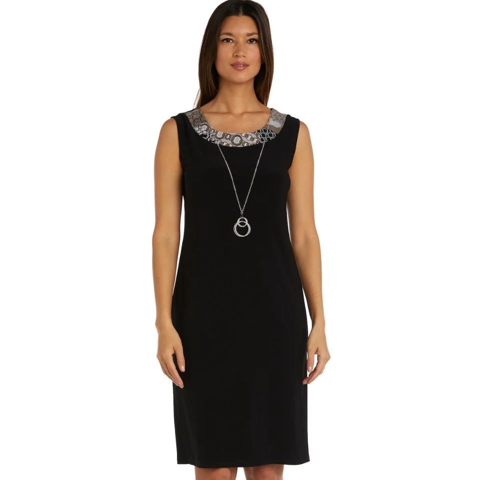 A woman modeling a sleeveless black knee-length dress with a decorative silver and black patterned yoke at the neckline, paired with a long silver pendant necklace and beige heeled sandals, against a plain white background.
