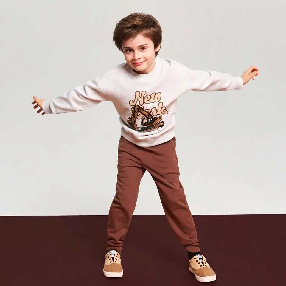 A young boy modeling a light gray sweatshirt with a brown graphic print and brown pants, posing with outstretched arms against a split white and dark brown studio background.