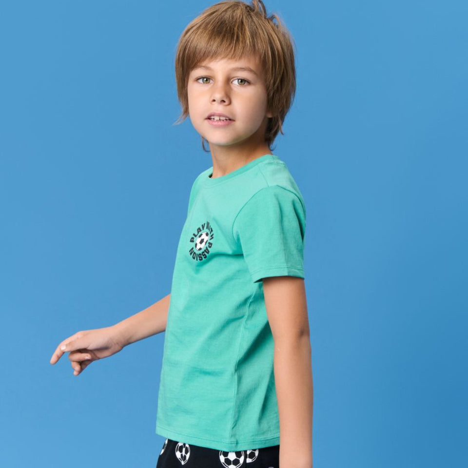 A young boy modeling a short-sleeve t-shirt in mint green with a soccer-themed graphic on the left chest, paired with black shorts featuring a white soccer ball pattern, set against a solid blue studio background.
