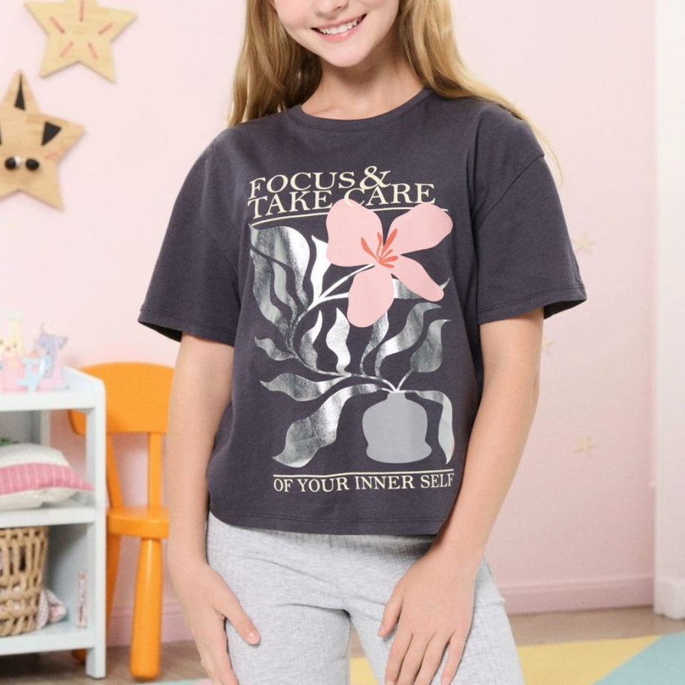 A young girl with blonde hair smiling, wearing a dark charcoal gray graphic t-shirt with a pink flower and the text 'FOCUS & TAKE CARE OF YOUR INNER SELF', paired with light gray sweatpants in a decorated children's room.