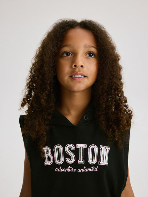 Young girl wearing a black sleeveless hoodie with 'Boston adventure unlimited' text on a plain background