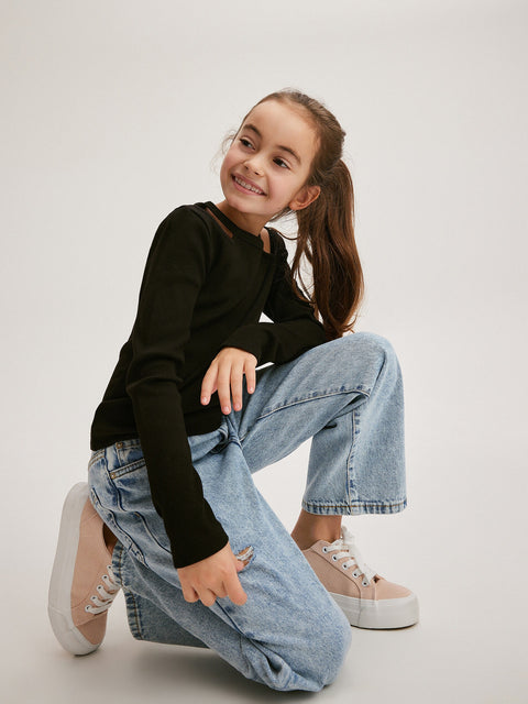Young girl wearing a black sweater and light blue jeans sitting on the floor against a white background