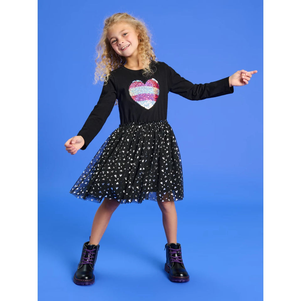 A young girl with curly blonde hair wearing a black long-sleeve dress with a sequined heart on the chest and a black tulle skirt with silver star patterns. She is wearing black boots with purple laces and posing against a solid blue studio background.