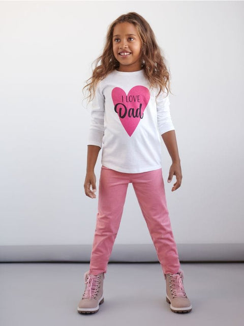 Young girl wearing a white shirt with a pink heart and text, pink pants, and pink shoes on a plain background