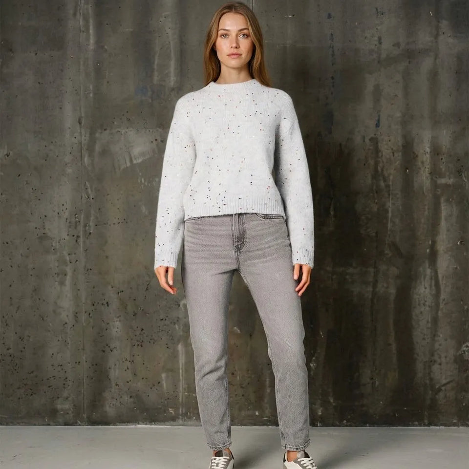 A woman stands against a textured concrete wall wearing a light gray speckled knit sweater, gray jeans, and white sneakers.