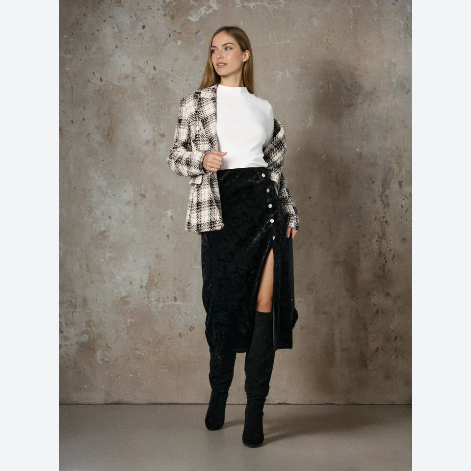 A woman stands against a textured concrete wall wearing a black and white plaid blazer over a white mock-neck top, a black velvet high-slit midi skirt, and black over-the-knee boots.