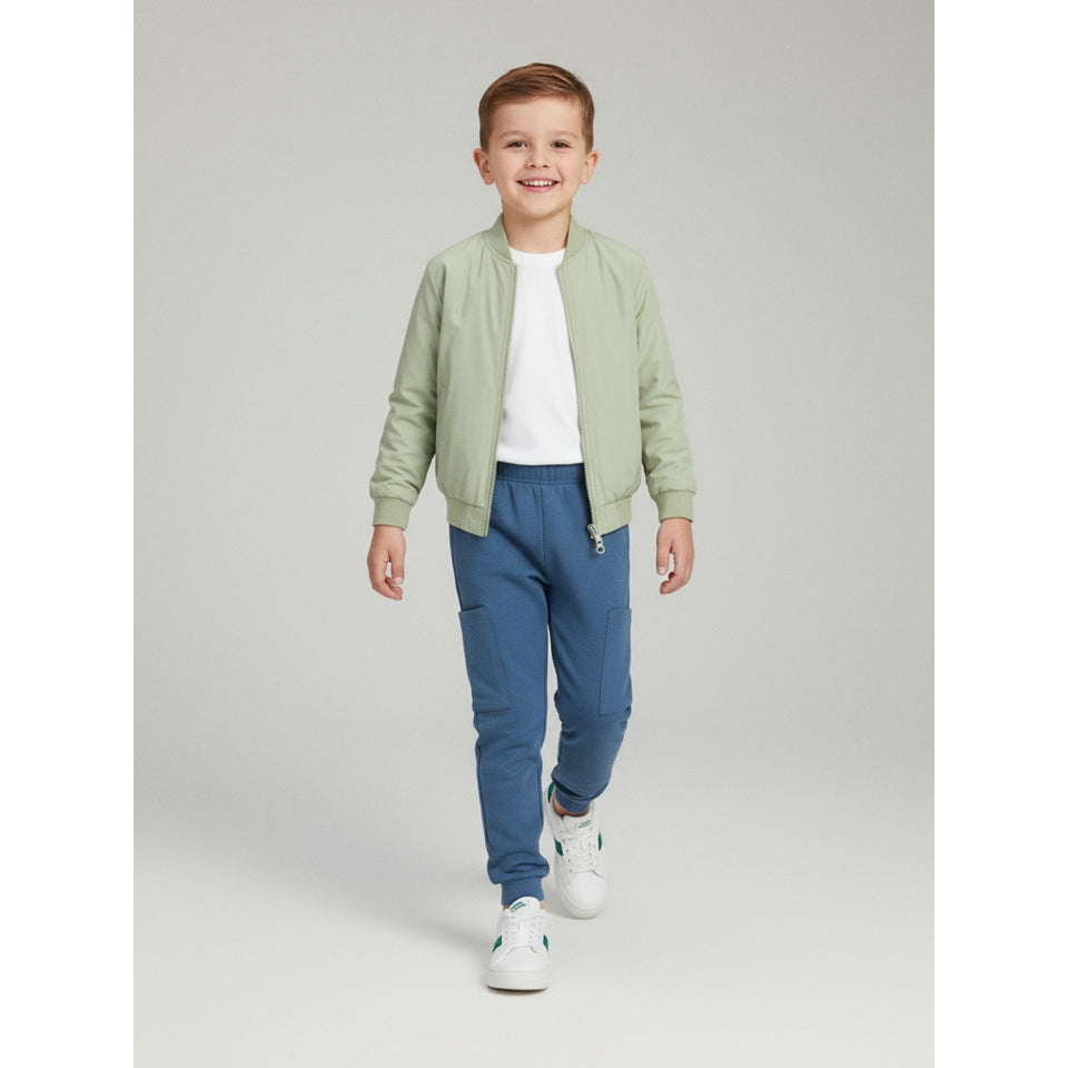 A young boy walking forward in a studio setting, wearing a light sage green zip-up bomber jacket over a white t-shirt, blue jogger pants, and white sneakers.