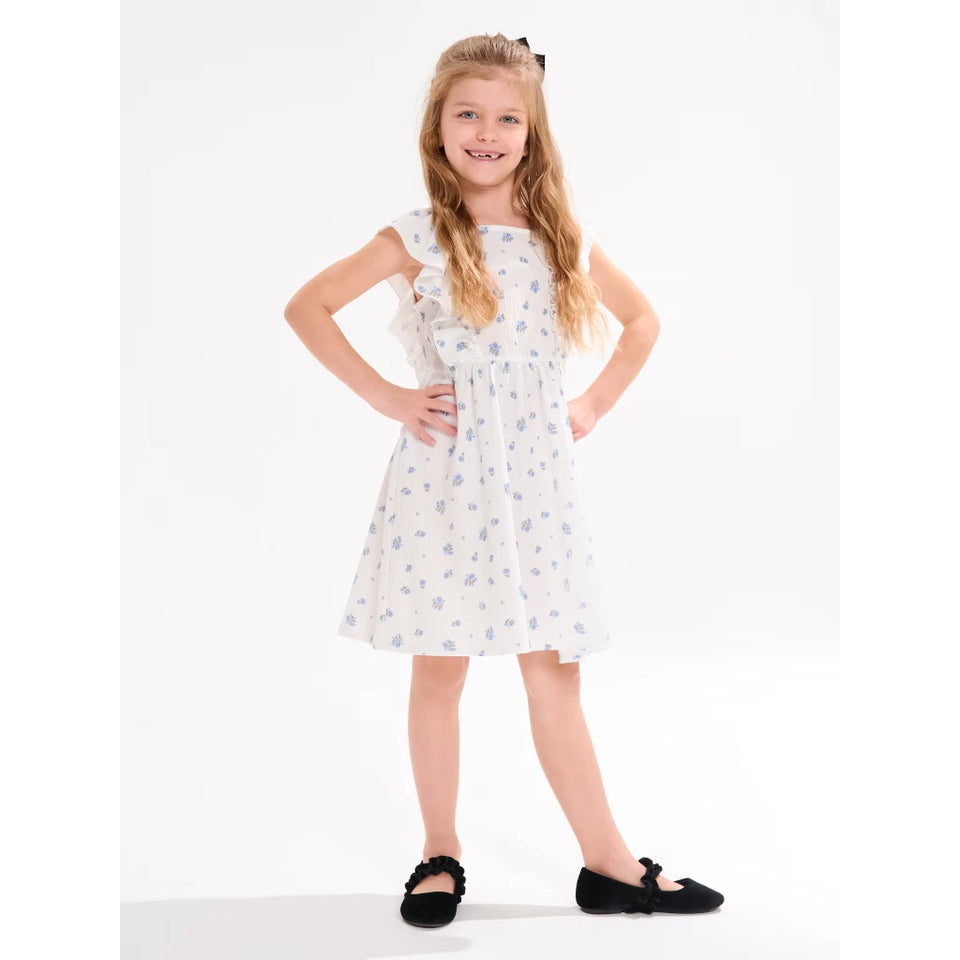 A young girl with blonde hair wearing a white sleeveless dress with a light blue floral pattern, standing with hands on hips against a plain white studio background.