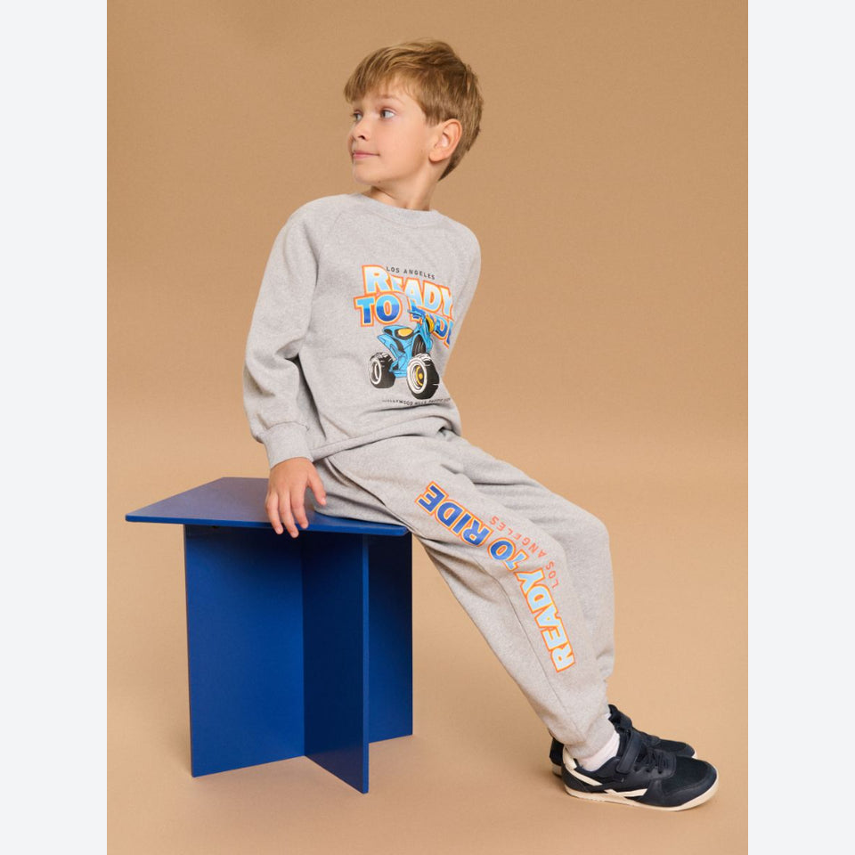 A young boy wearing a light gray two-piece sweatshirt and jogger set with a blue and orange 'Ready to Ride' graphic, sitting on a blue block against a solid tan background.