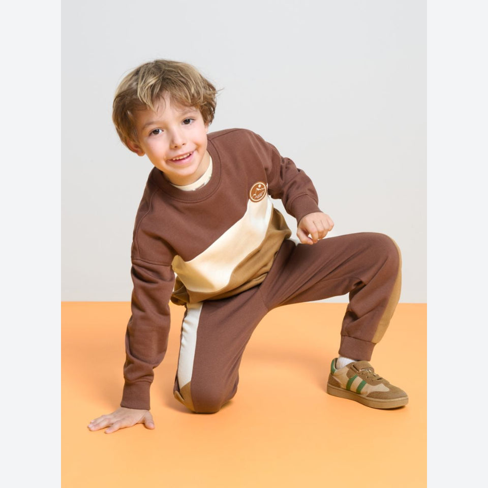 A young boy wearing a brown and beige two-piece sweatshirt and jogger set, posing on an orange floor against a light gray background.
