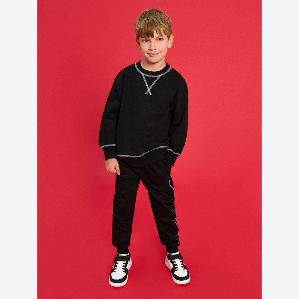 A young boy modeling a black crewneck sweatshirt and matching black track pants with silver contrast stitching, paired with black and white sneakers, set against a solid red studio background.