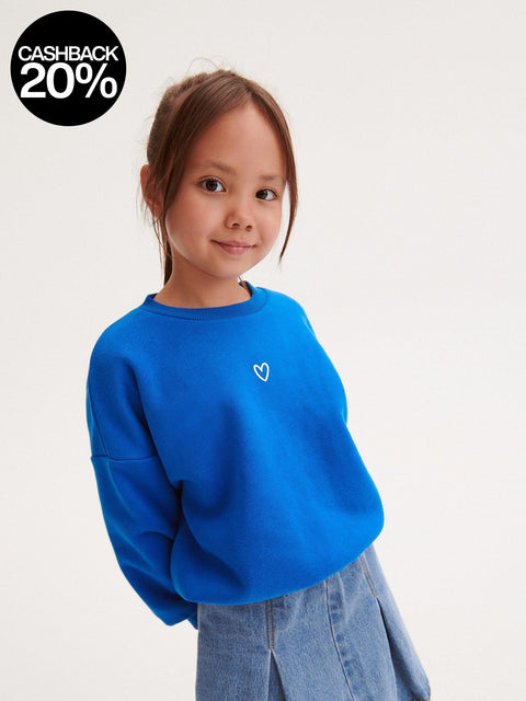 Young girl wearing a blue sweatshirt with a white heart on a plain background